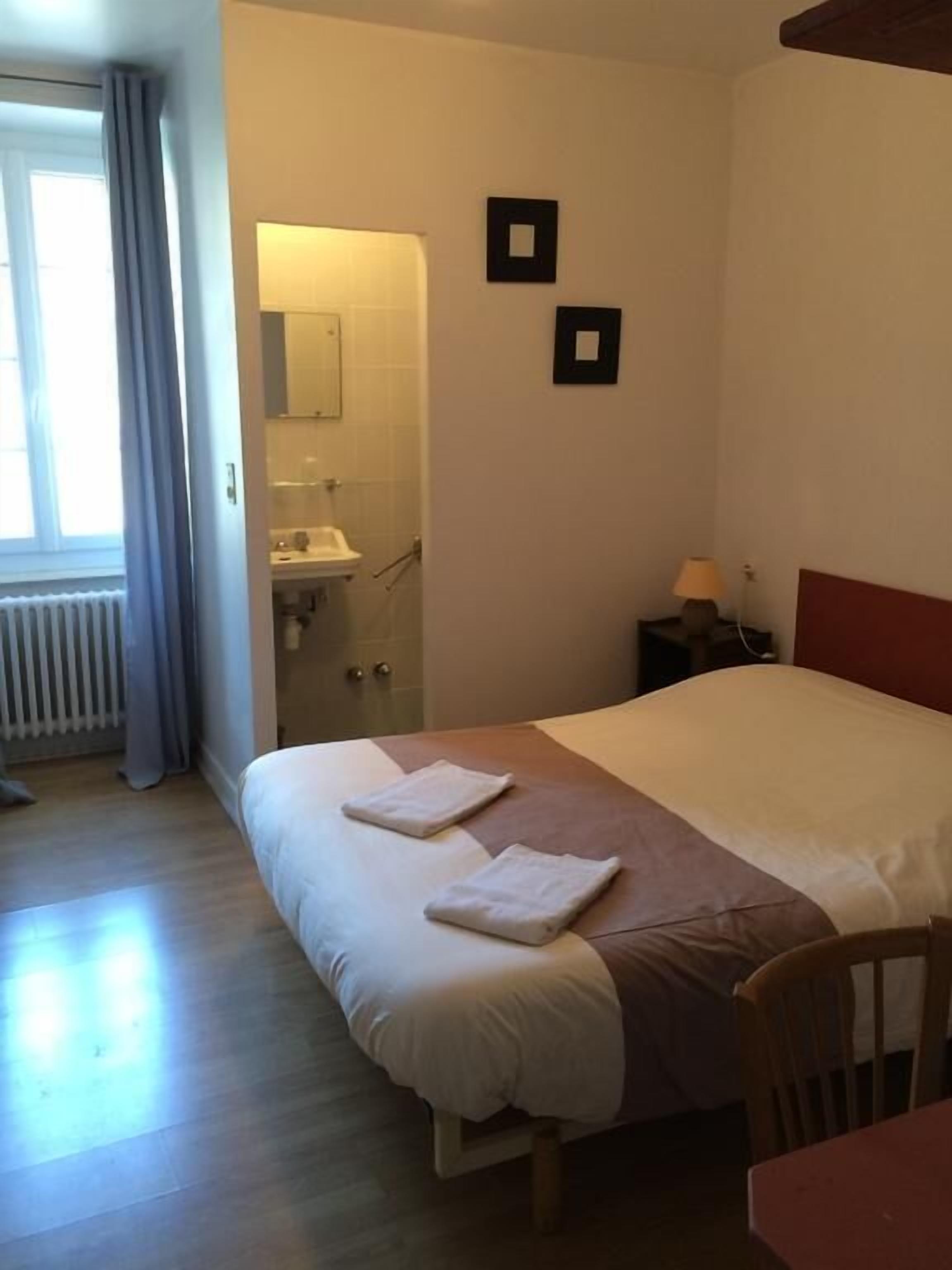 Quarto de hotel com cama, cadeira de madeira, mesa de cabeceira, luminária, janela com cortinas e banheiro visível por uma porta aberta.