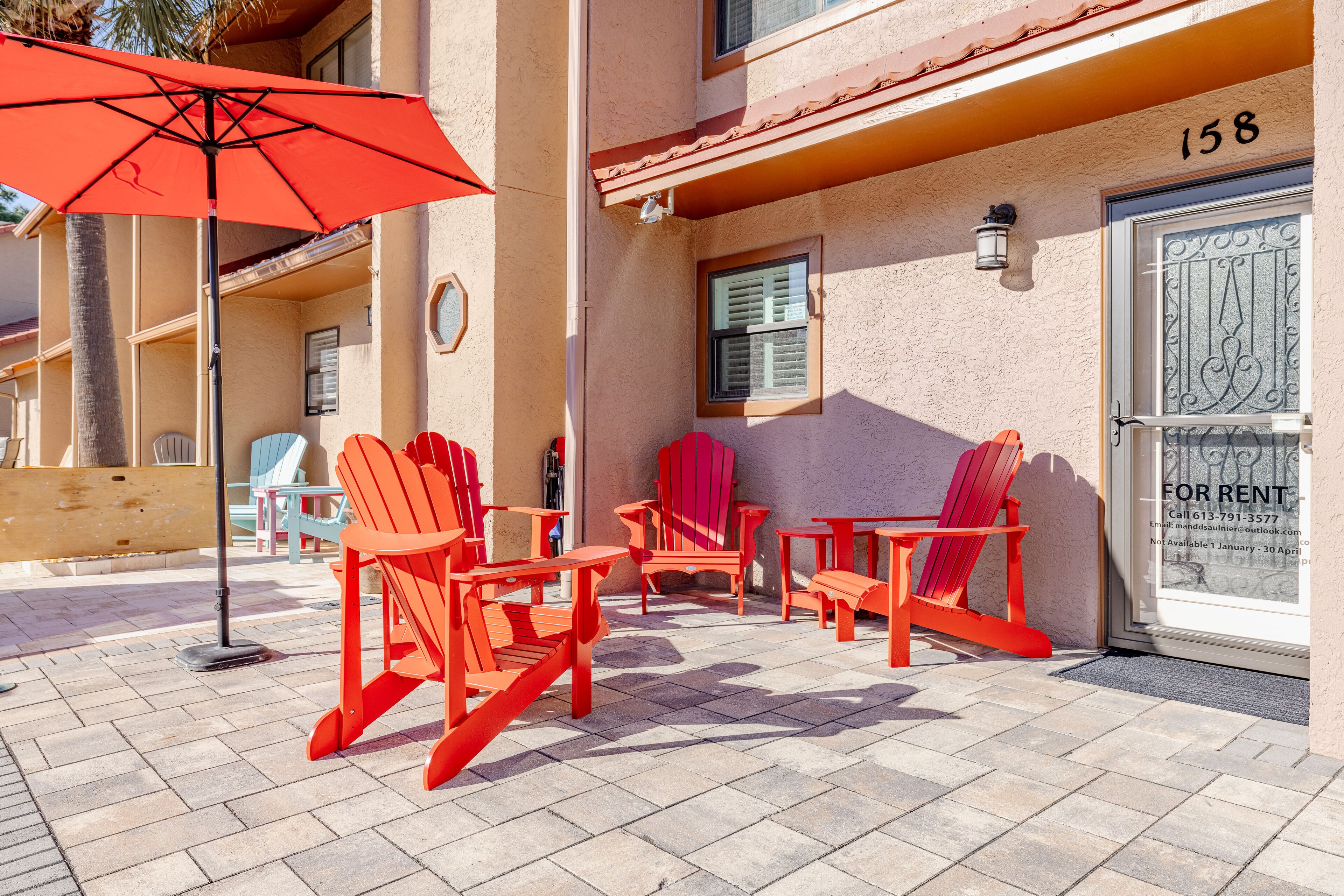 Eine Terrasse mit roten Adirondack-Stühlen, einem roten Sonnenschirm und einem Schild mit der Aufschrift 'For Rent' am Gebäude.