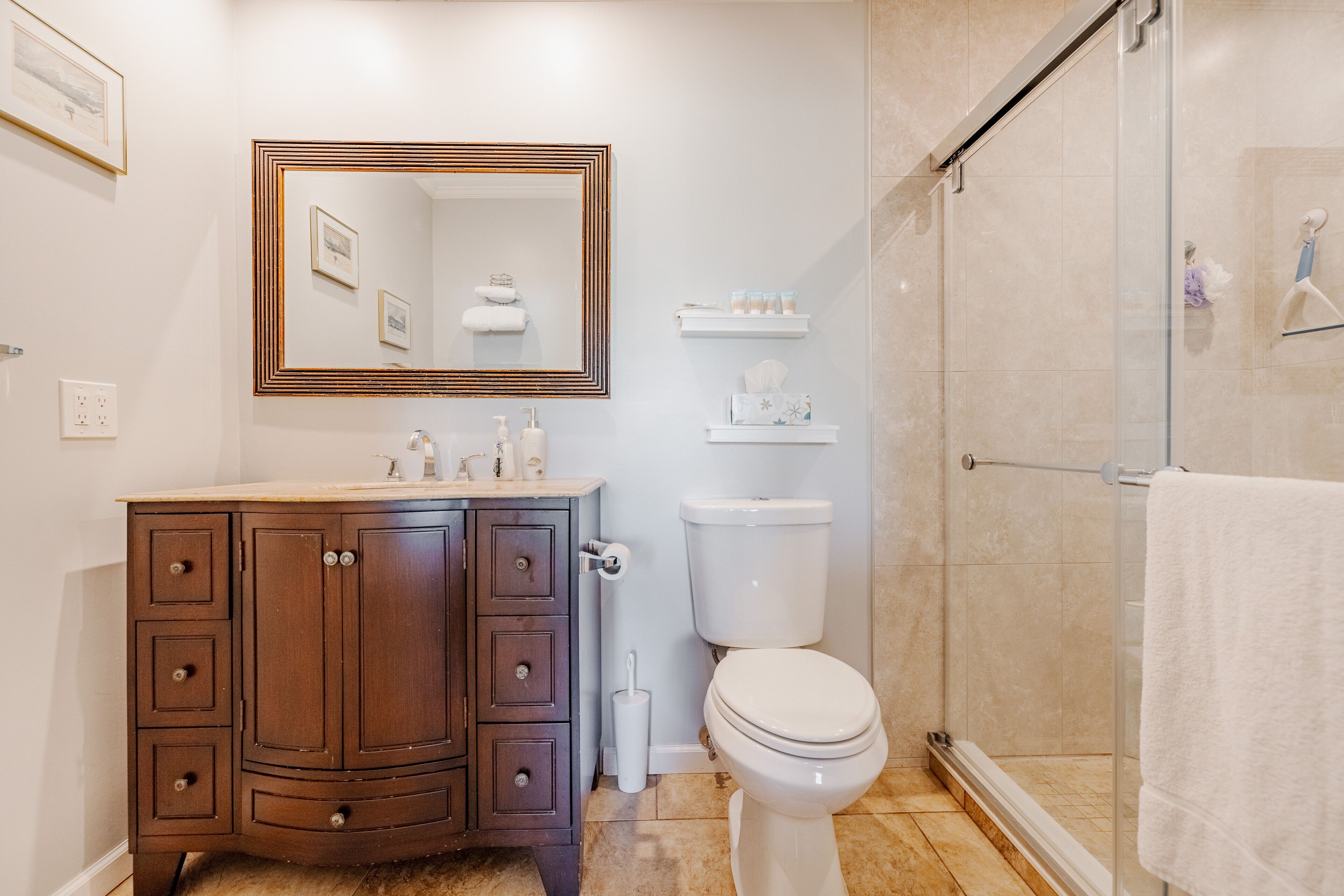A bathroom with a wooden vanity, a toilet, a shower with a glass door, and a mirror with a decorative frame.