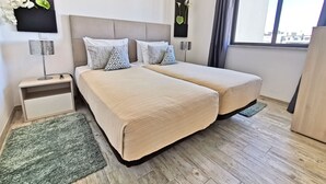 Une chambre d’hôtel moderne avec deux lits, une table de chevet, une lampe, un téléviseur fixé au mur et une fenêtre avec des rideaux.