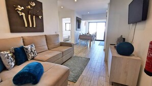 Un salon moderne avec un canapé beige, un coussin bleu, un téléviseur à écran plat fixé au mur et un coin repas à l’arrière-plan.
