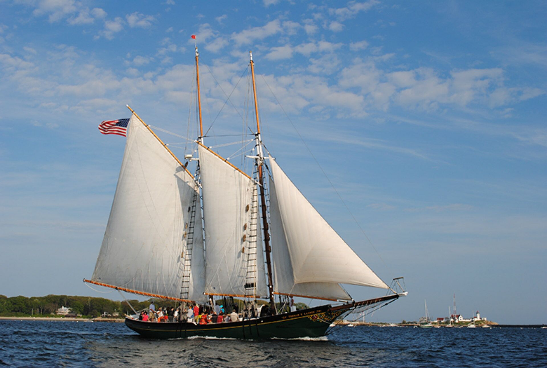 A sailboat with multiple sails and a flag, sailing on water with a distant shoreline and buildings.