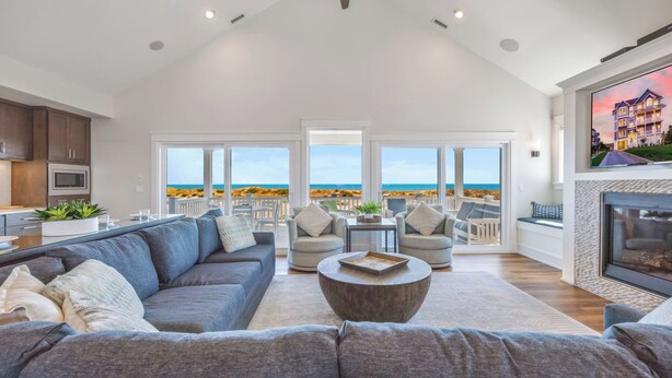 Ett rymligt vardagsrum med en stor grå soffa, ett soffbord och utsikt över stranden genom stora fönster.