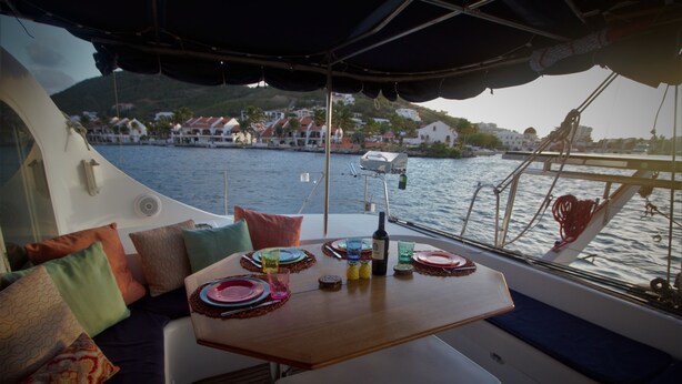 A boat's interior with a dining table set for four, surrounded by colorful cushions and a view of a coastal town. - Dreamcatcher (Cole Bay)