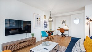 Een moderne woonkamer met een flatscreen televisie, een salontafel en een bank. Er is een eethoek met een tafel en stoelen, en een klein bureau met een lamp.