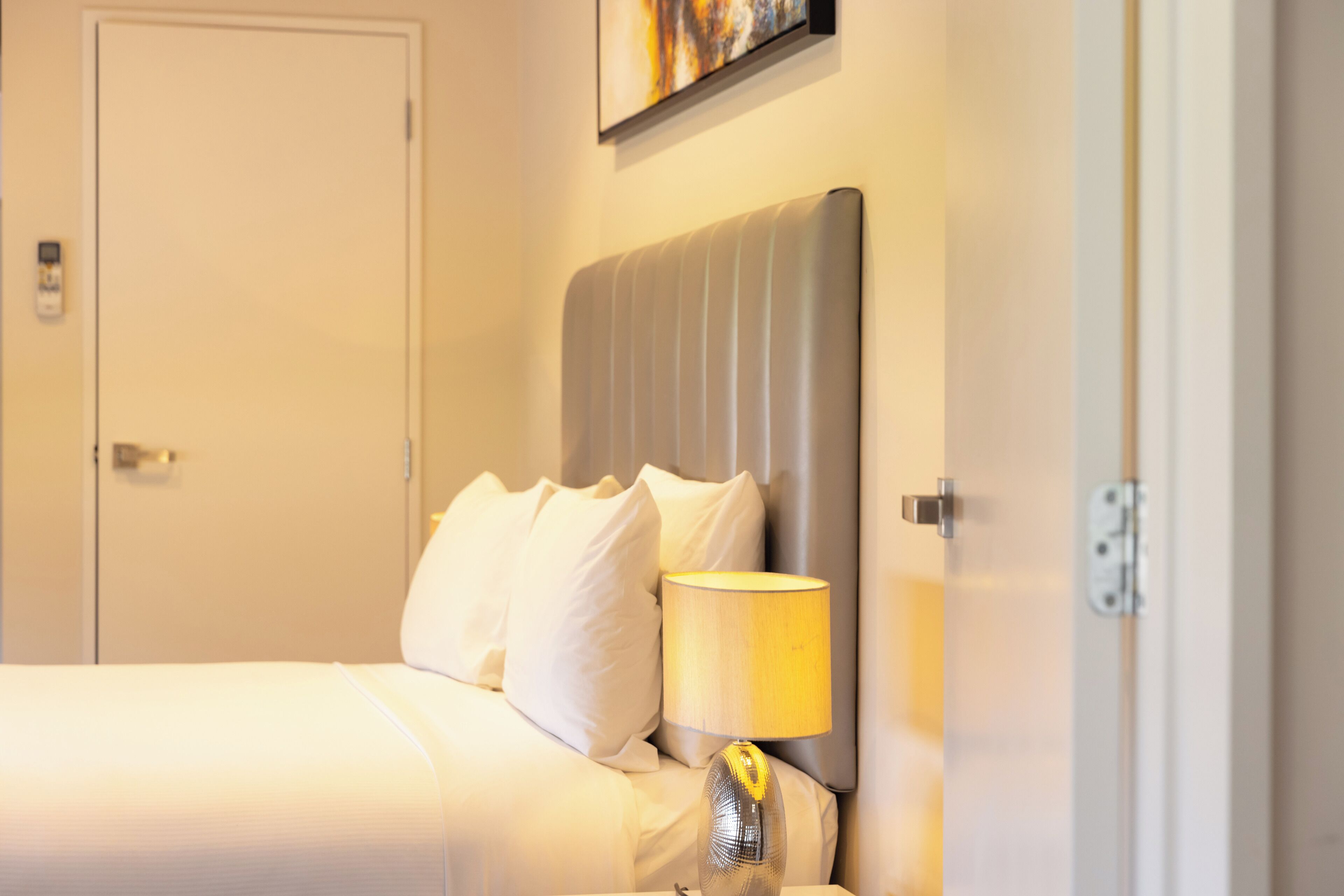 Quarto de hotel com cama, mesa de cabeceira e uma luminária, e uma porta.