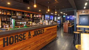 A bar area with a wooden counter labeled "HORSE & JOCKEY," shelves with bottles and glasses, and a seating area with stools. - Alpine Hotel (Christchurch)