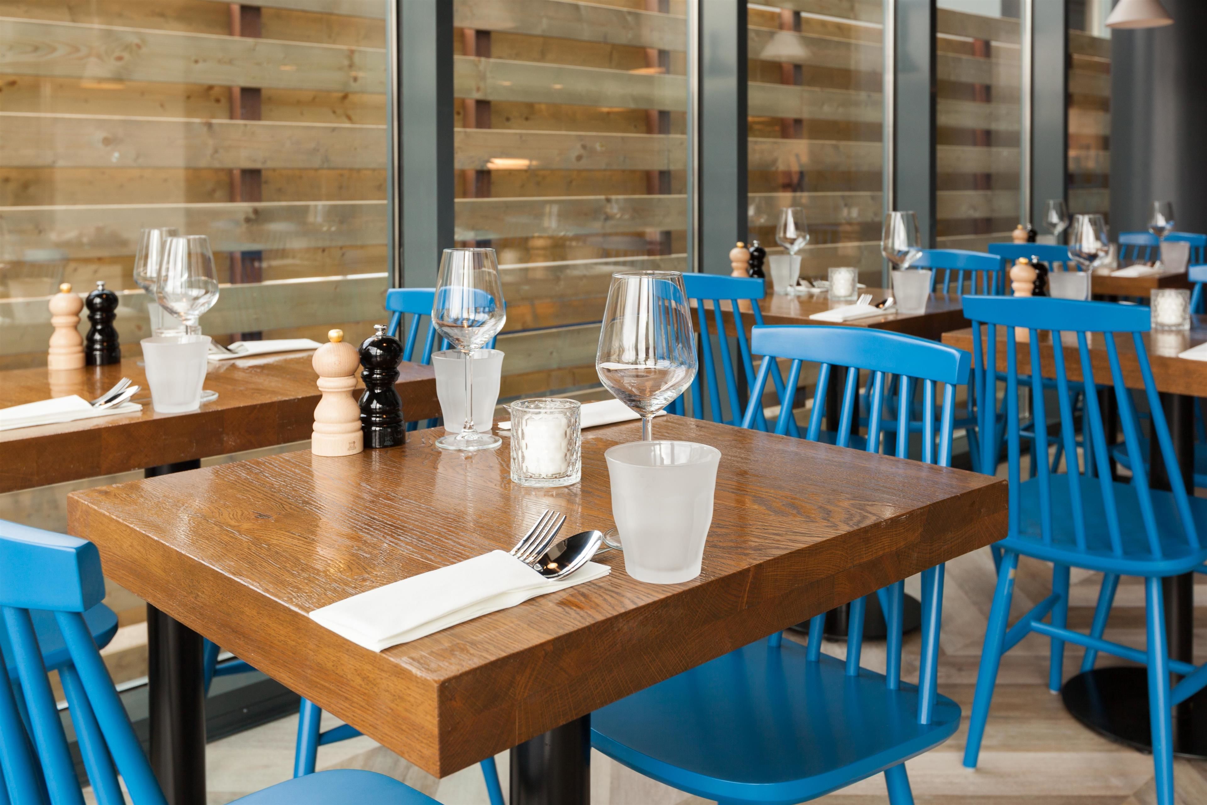 Interior de restaurante com mesas de madeira e cadeiras azuis, copos de vidro transparentes e saleiro e pimenteiro nas mesas.