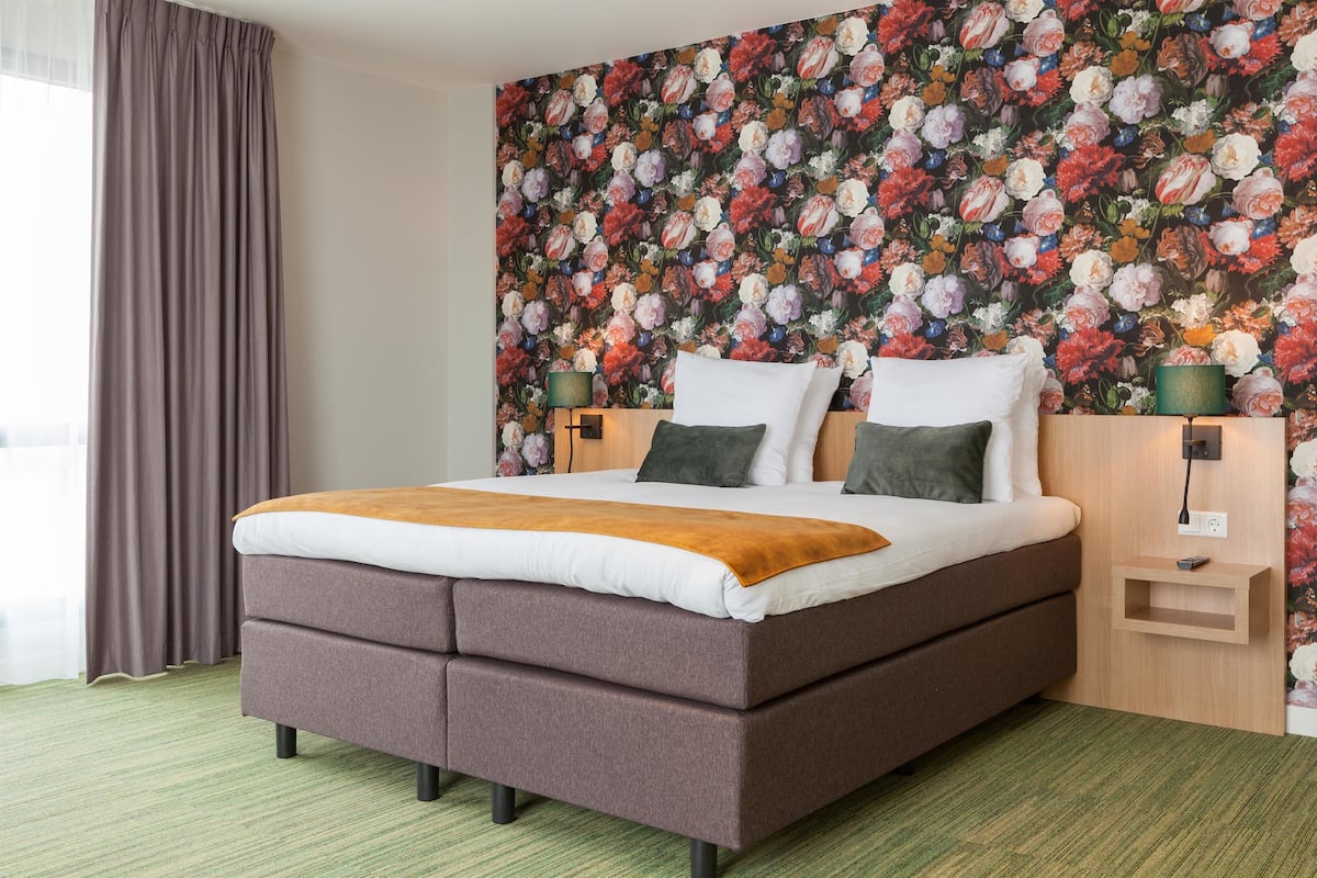 Camera d'albergo moderna con un letto grande, carta da parati floreale, un comodino in legno e un tappeto verde.