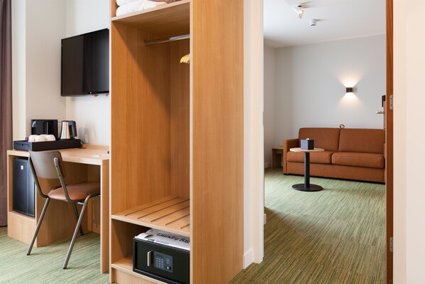 Habitación de hotel pequeña con un armario de madera, un escritorio con silla, televisor de pantalla plana, sofá y una mesita.