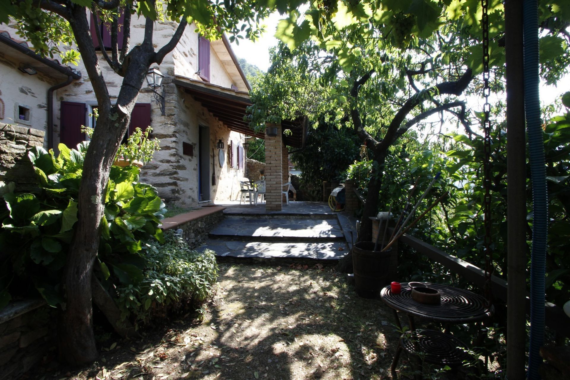 A stone building with a covered porch, surrounded by lush greenery and a garden area.
