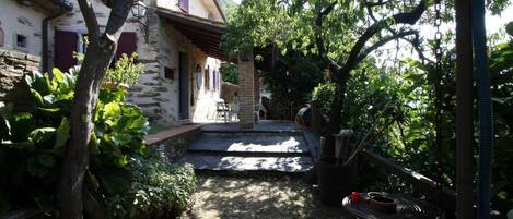 A stone building with a covered porch, surrounded by lush greenery and a garden area.