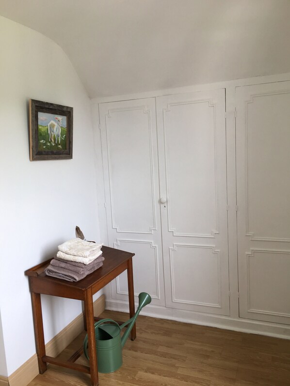 Quarto com mesa de madeira, cobertores dobrados, um regador verde e um quadro na parede.