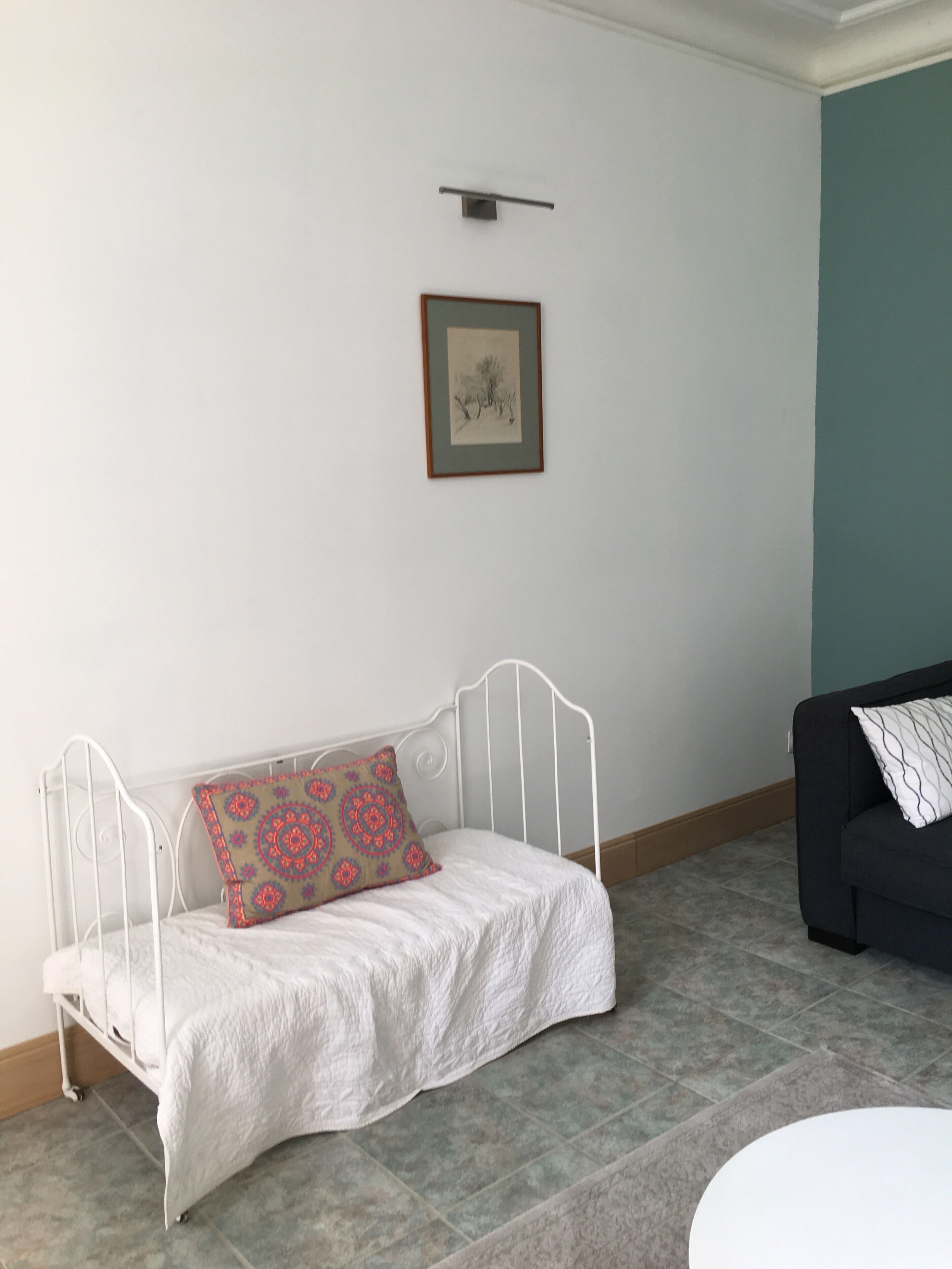 A white metal bed with a patterned pillow, a framed picture on the wall, and a dark sofa in a room with tiled flooring.