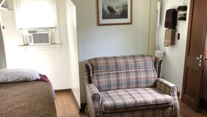 Una habitación con dos camas, un sofá a cuadros, un cuadro enmarcado en la pared y una ventana con persianas.