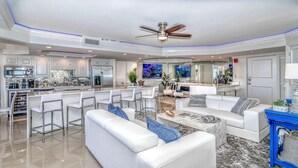 A modern living room with a white sofa, a coffee table, and a TV. There is a kitchen area with a microwave and a refrigerator.