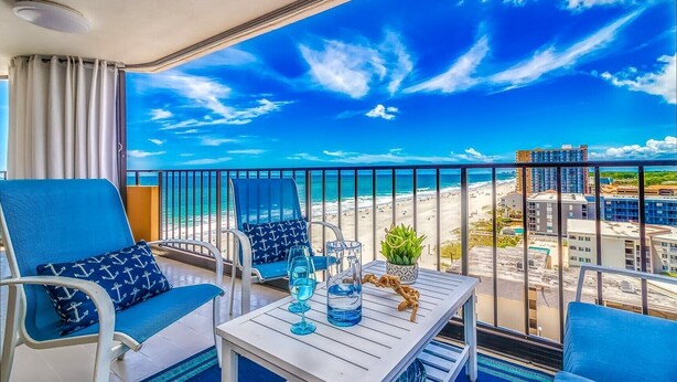 A balcony with a view of the beach and cityscape, furnished with blue chairs and a table.