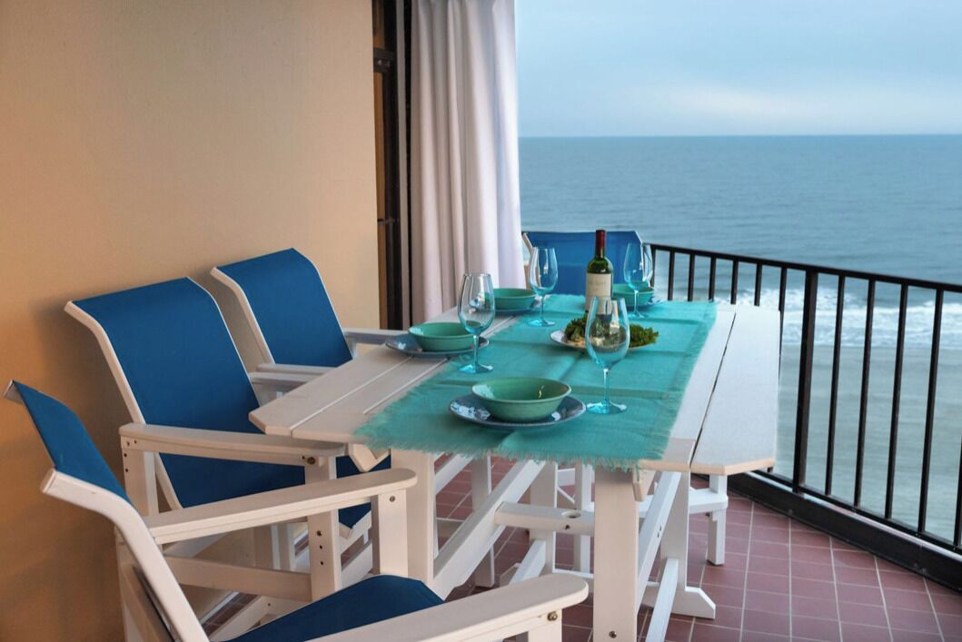A balcony table set with blue chairs, a tablecloth, and a view of the ocean.