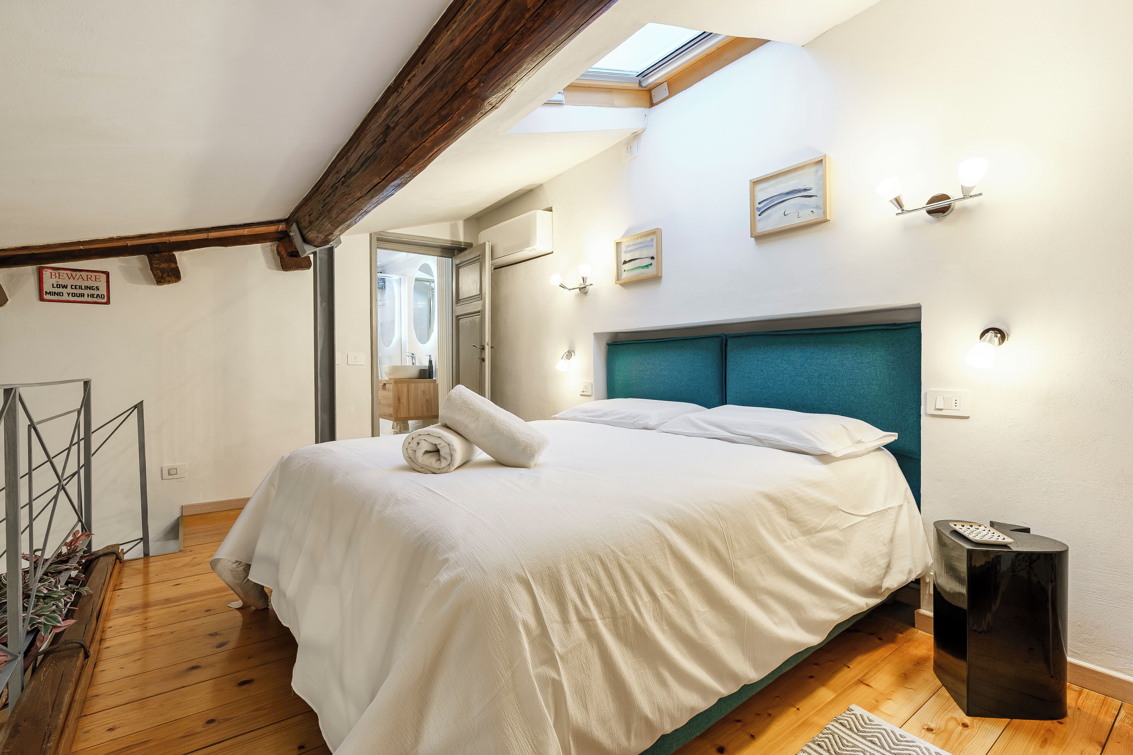 Een slaapkamer met een groot bed, houten balken, een dakraam, een nachtkastje en een wandlamp.