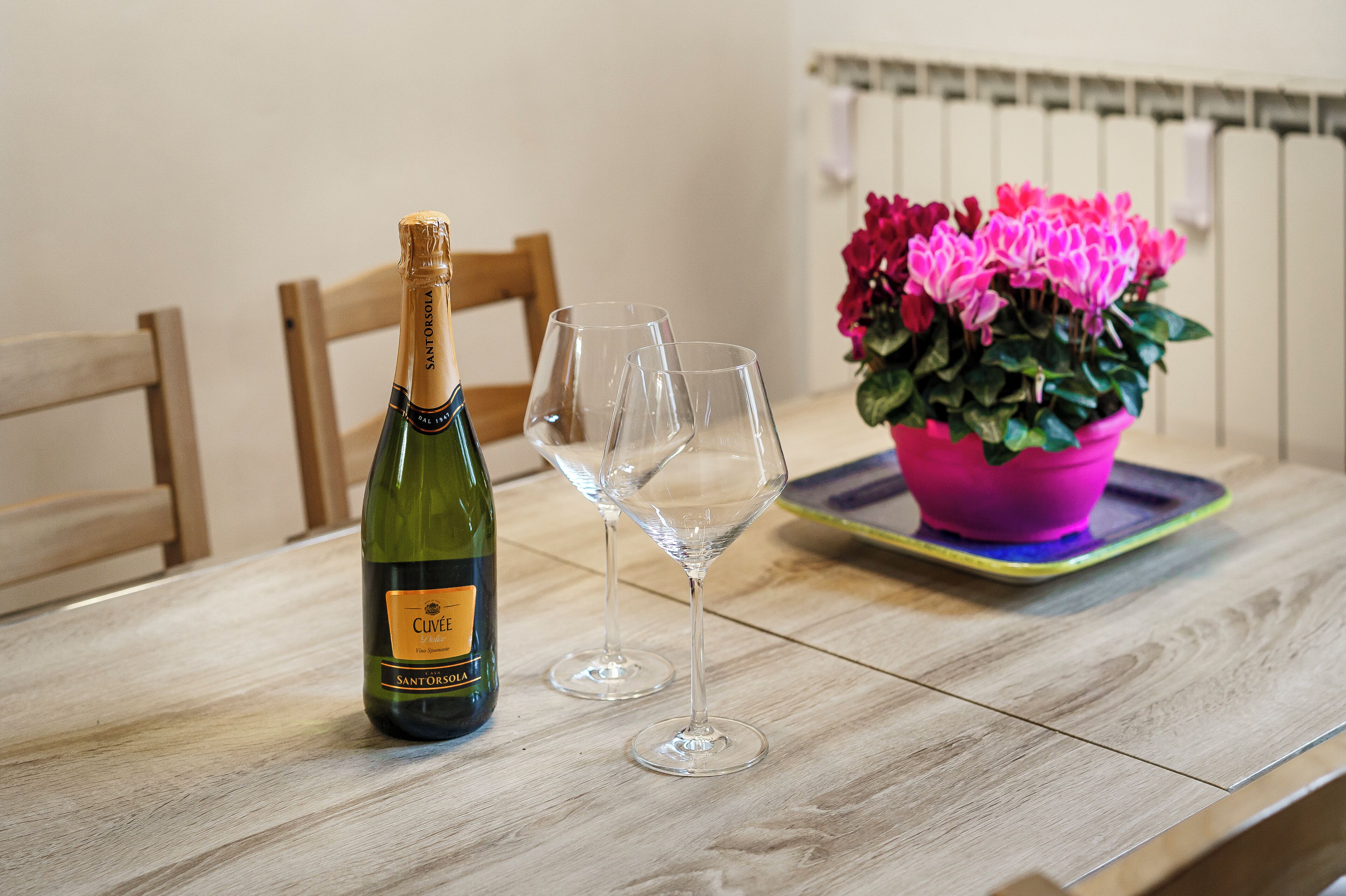 A wooden table with a bottle of Clive wine, two empty wine glasses, and a potted plant with pink flowers.