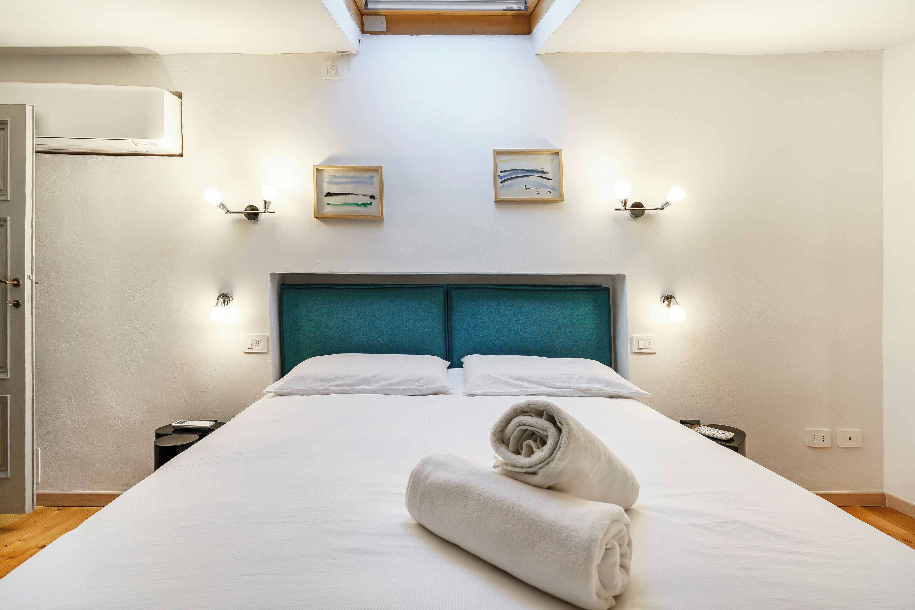 A hotel room with a large bed, a teal headboard, two wall-mounted light fixtures, and a skylight above.