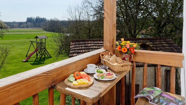 Ein Balkon mit Holzgeländer, ein Tisch mit Speisen und Getränken, eine Rutsche im Hintergrund und Blick auf ein grünes Feld.