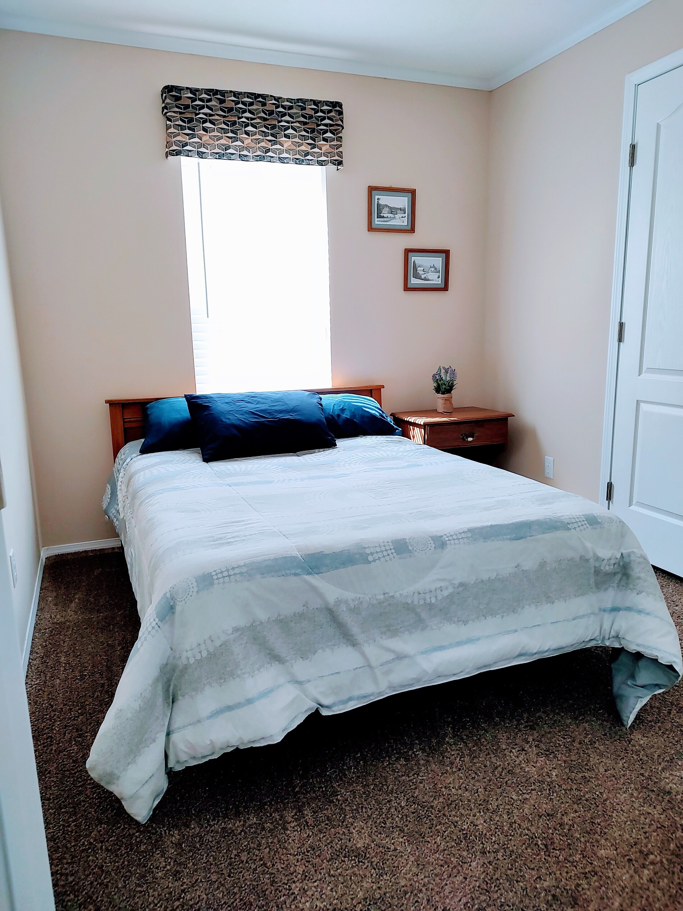 A bedroom with a bed, a nightstand, a window with patterned curtains, and two framed pictures on the wall.