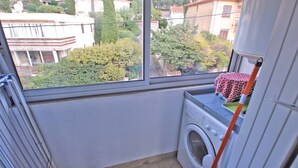 Un espacio para lavandería con lavadora y un tendedero, una ventana con vistas a edificios y árboles, y una mesa con mantel estampado.