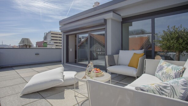 Une terrasse sur le toit dotée d’un espace salon extérieur moderne, comprenant un canapé et des fauteuils, ainsi qu’une petite table sur laquelle est posé un panier de fruits.