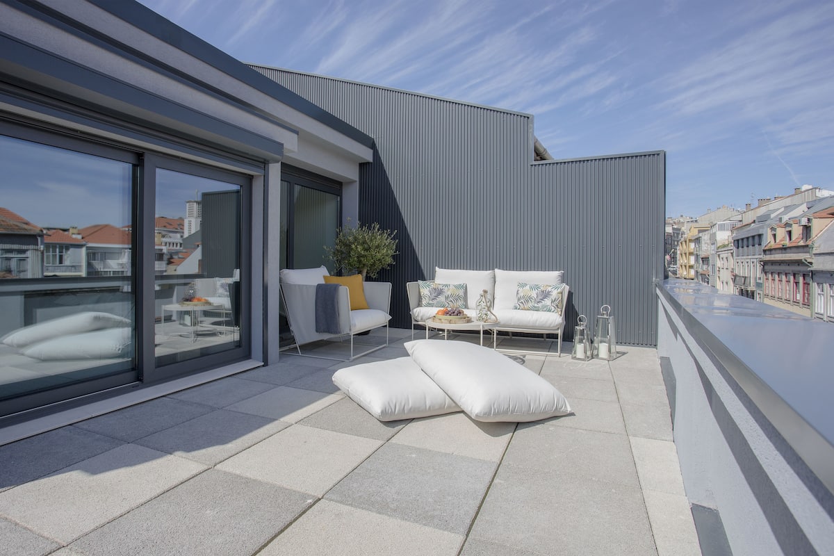 Un área de estar moderna al aire libre con un sofá, cojines y una pequeña mesa, con vistas a un paisaje urbano.