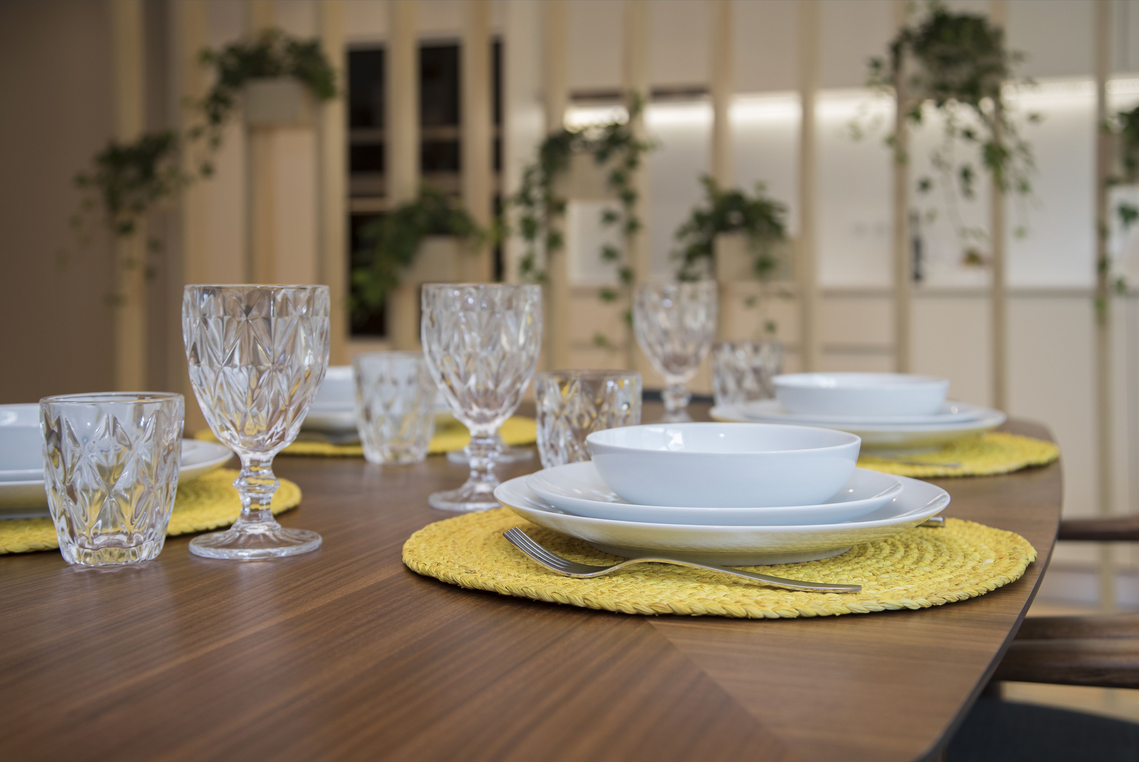 A dining table set with clear glassware and white plates, with a woven placemat and a fork on it.