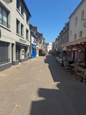 Une rue bordée d’immeubles, un café avec une terrasse, et un ciel bleu dégagé.