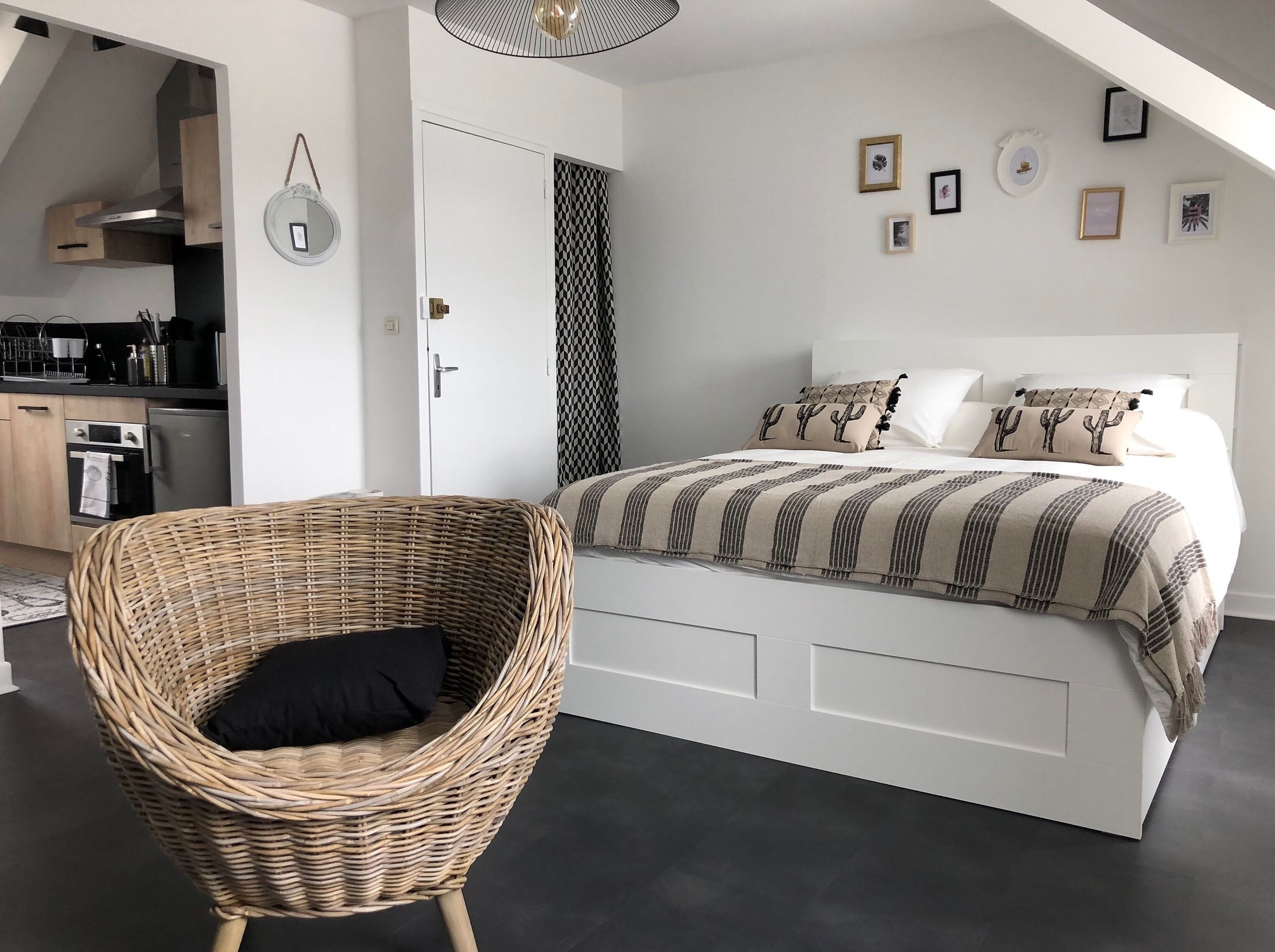 A bedroom with a bed, a wicker chair, a wardrobe, and a kitchen area.