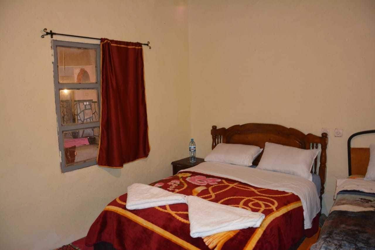 Un dormitorio con una cama de madera, una ventana con cortina, una mesita de noche con una botella de agua y una colcha con toallas.
