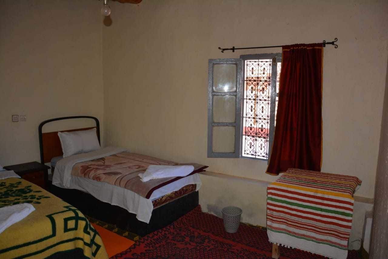 A small, simple room with a single bed, a window with a metal grill, and a red curtain.