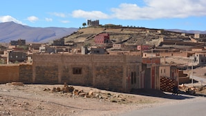 Un poble rural amb edificis de pedra tradicionals, una estructura vermella destacada en un turó i un camí al primer pla.