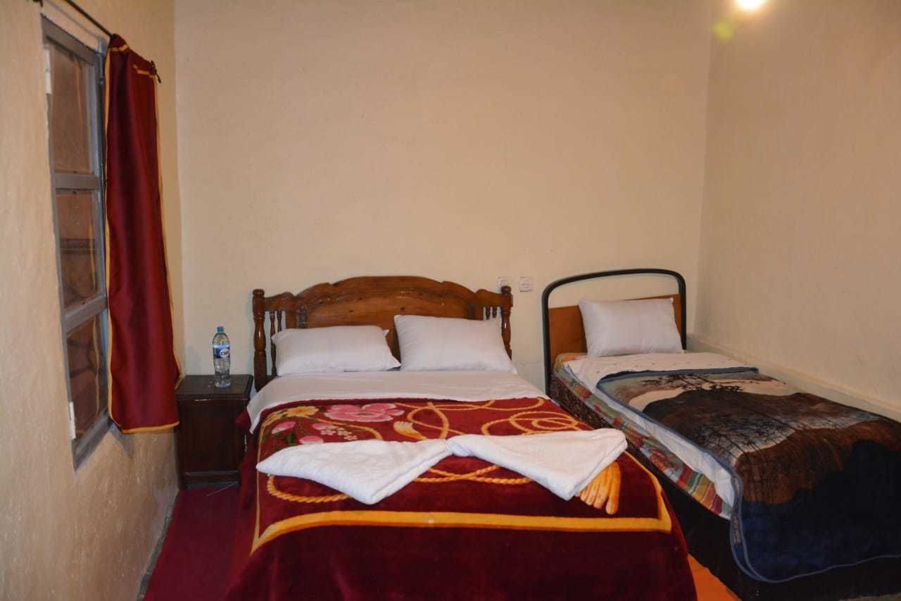 A small, single-room hotel room with two beds, a wooden headboard, a nightstand, and a window with a red curtain.