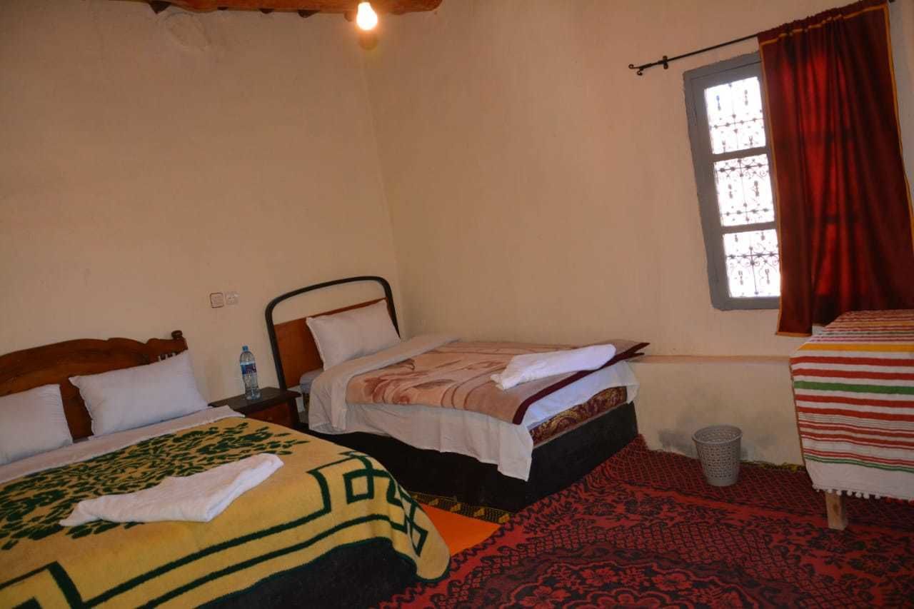 A room with two beds, a window with red curtains, a wooden ceiling light, and a patterned rug.