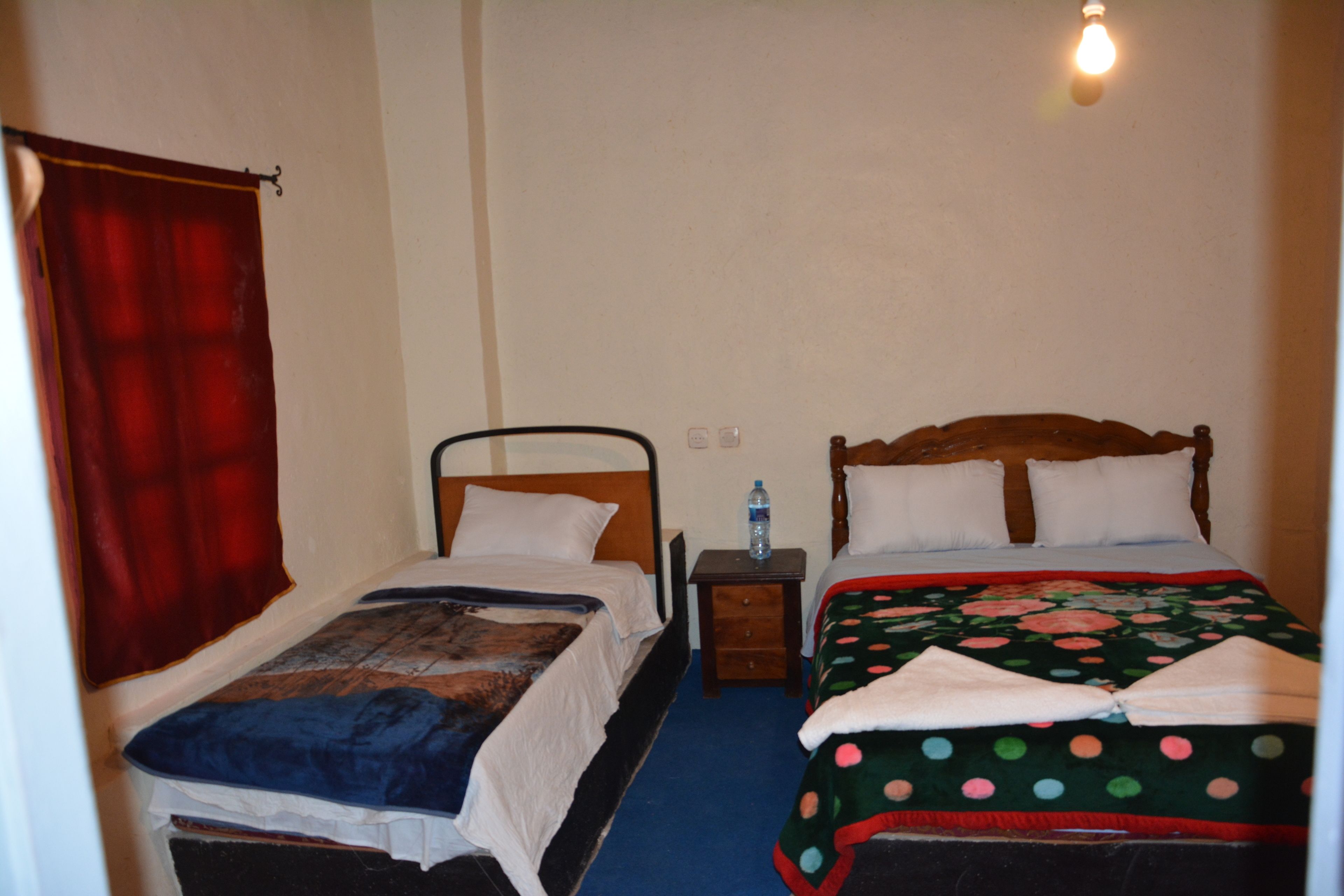 A room with two single beds, a wooden headboard, a nightstand, a water bottle, and a red curtain.
