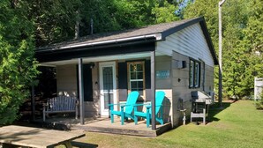 Eine kleine Holzhütte mit Veranda, zwei türkisfarbenen Adirondack-Stühlen, einem Picknicktisch und einem Schild mit der Aufschrift 'Cedar Cove Cabin'.