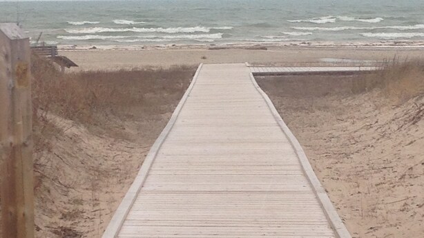 Ein Holzsteg führt zu einem Strand, im Hintergrund sind Wellen zu sehen.