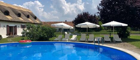 A swimming pool with lounge chairs and umbrellas, a thatched-roof building, and a grassy area with trees.