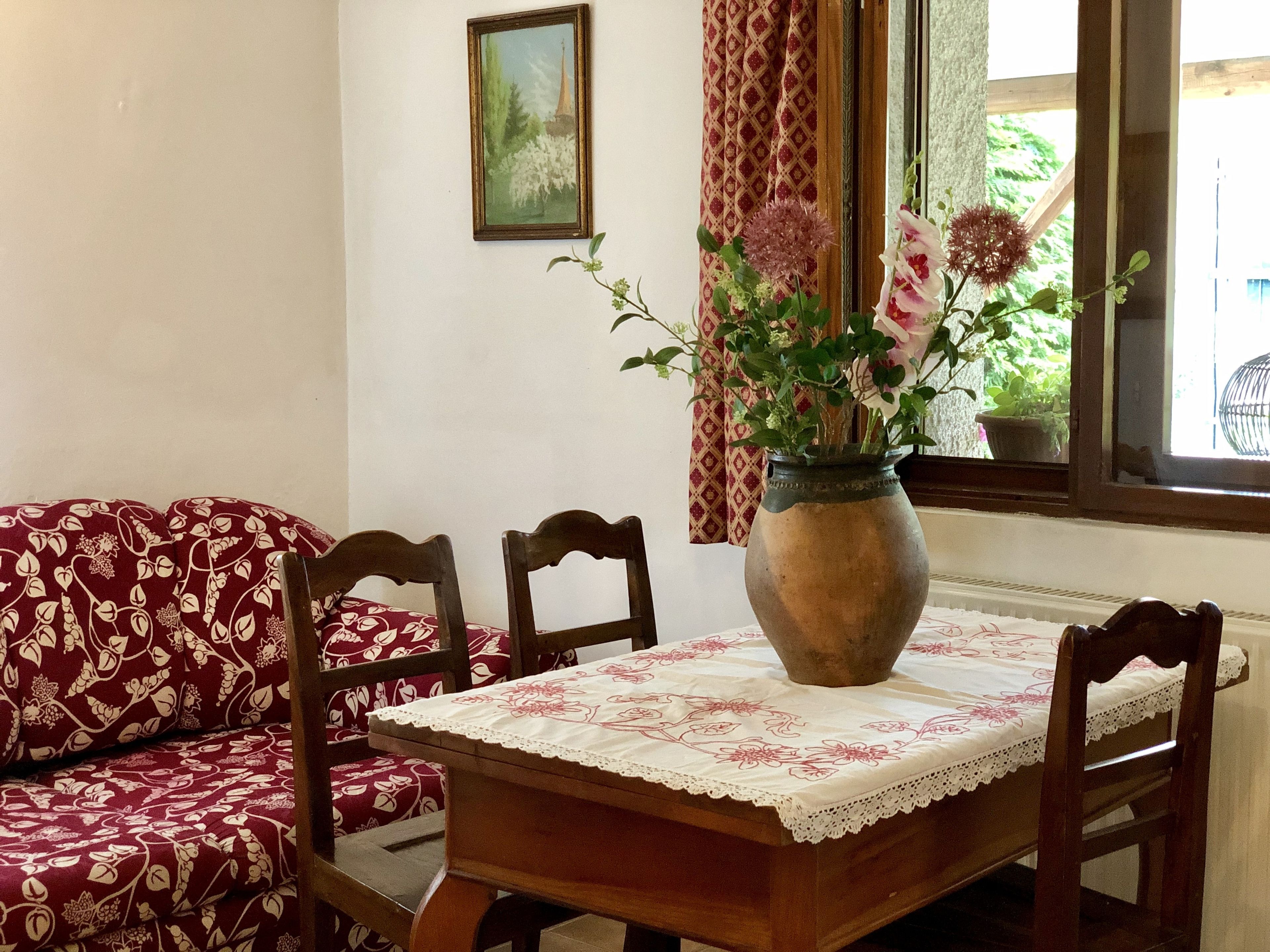 A dining area with a wooden table, chairs, a vase with flowers, a patterned sofa, a window with curtains, and a framed picture on the wall.