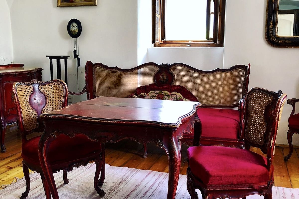 Une pièce au décor traditionnel, avec une table à manger en bois, des chaises à revêtement rouge, un canapé en osier, une horloge murale et un miroir.