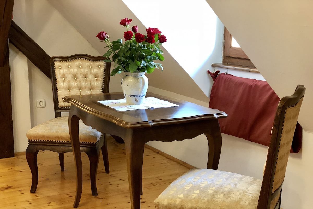 Un coin repas avec une table et des chaises en bois, un vase rempli de roses rouges et une vue sur le ciel grâce à une lucarne.