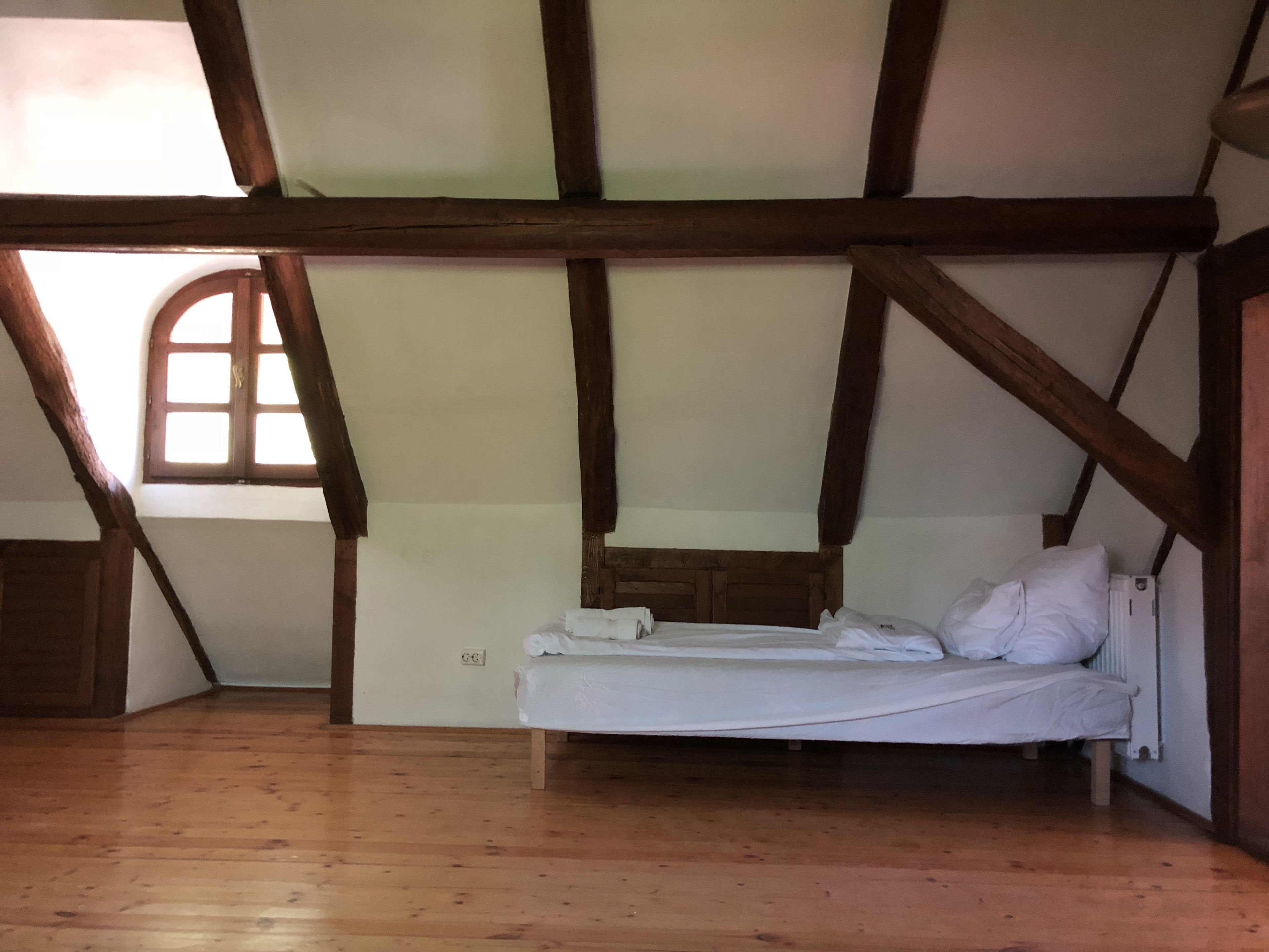 Une pièce avec un plancher en bois, un lit avec une literie blanche et une fenêtre avec un cadre en bois.