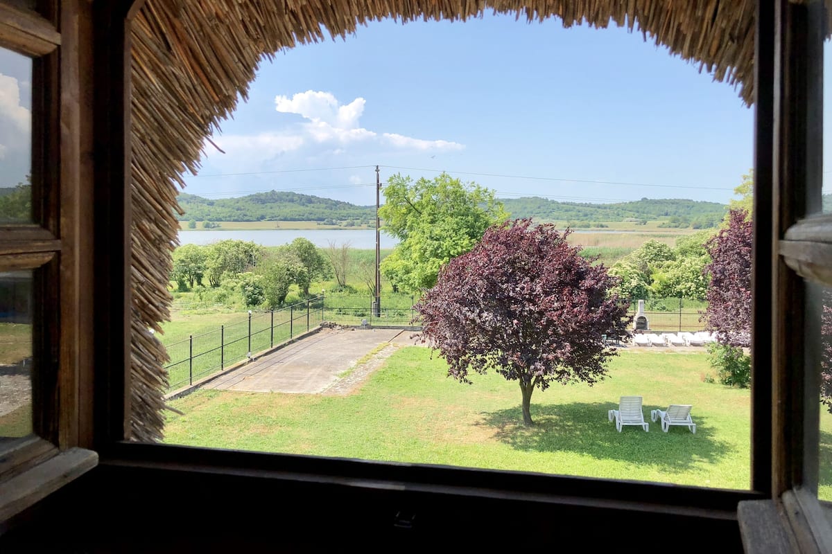 Une vue depuis une fenêtre surmontée d’un toit de chaume, donnant sur une zone herbeuse avec un arbre, deux chaises blanches et une étendue d’eau, avec des collines visibles au loin.