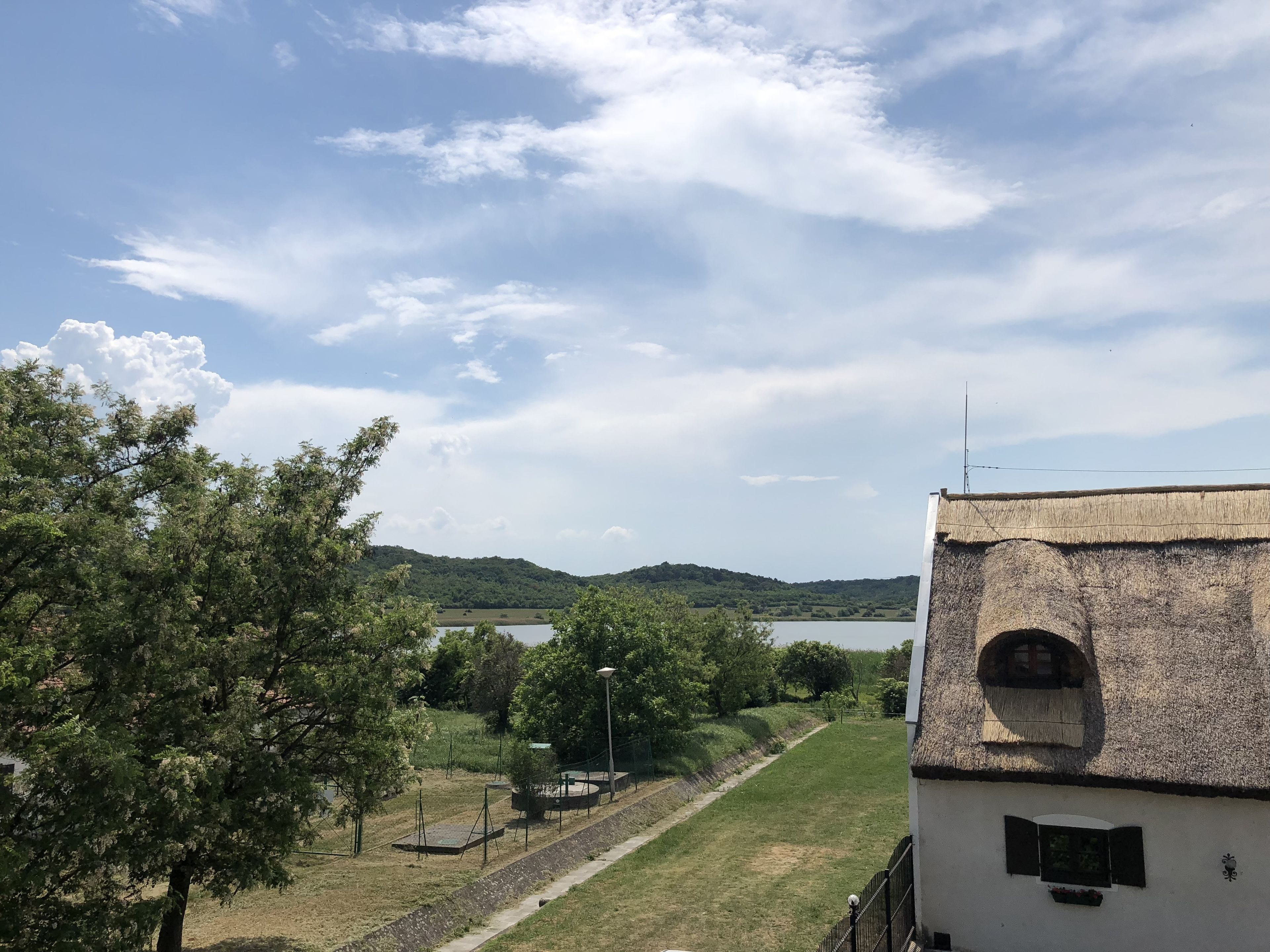 Blick auf ein Gebäude mit Strohdach und einem kleinen Fenster, mit Blick auf eine grüne Wiese mit Bäumen und einem entfernten Gewässer.