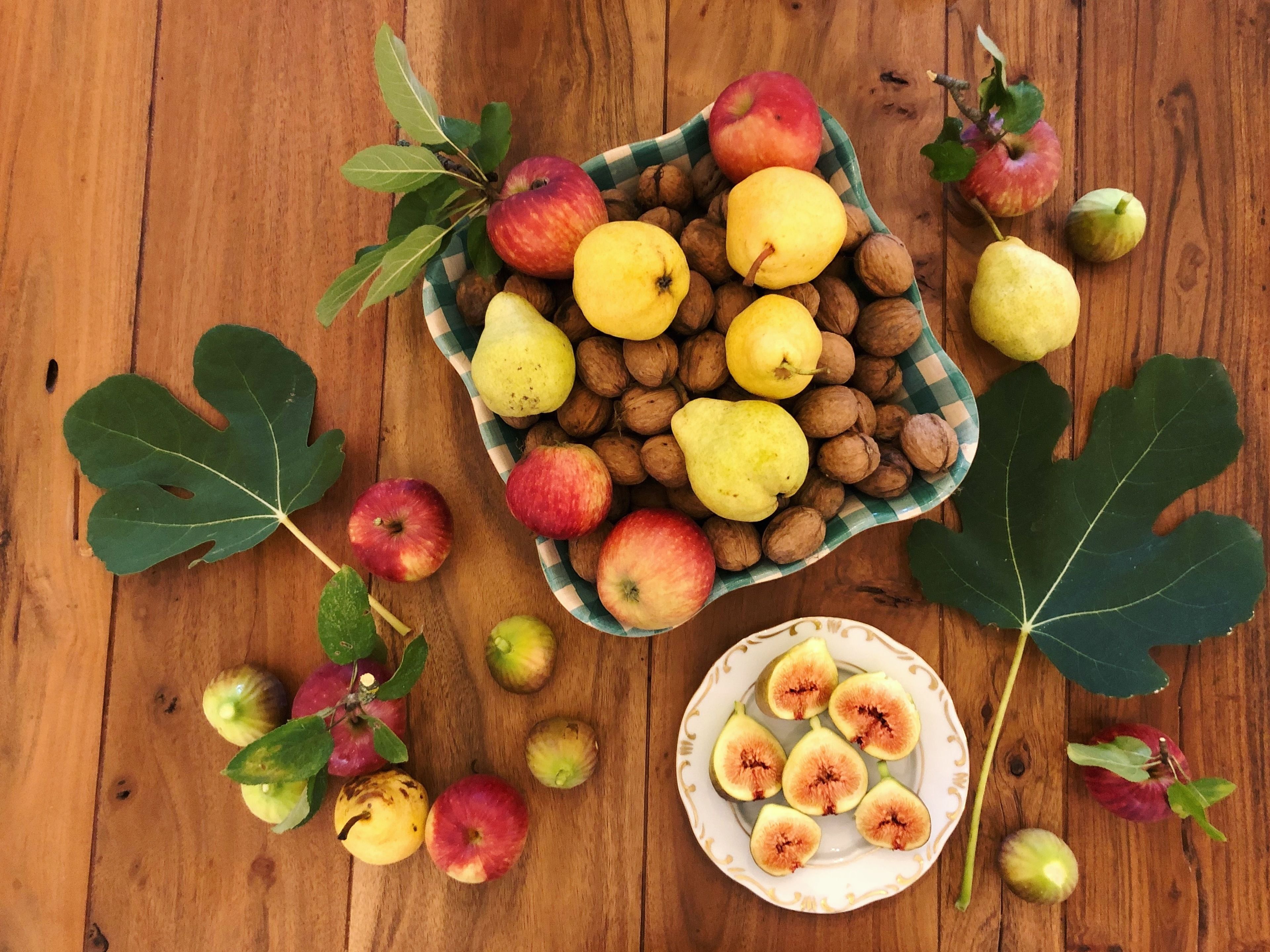 A wooden table with a bowl of fruits and nuts, a plate of sliced figs, and two large leaves.
