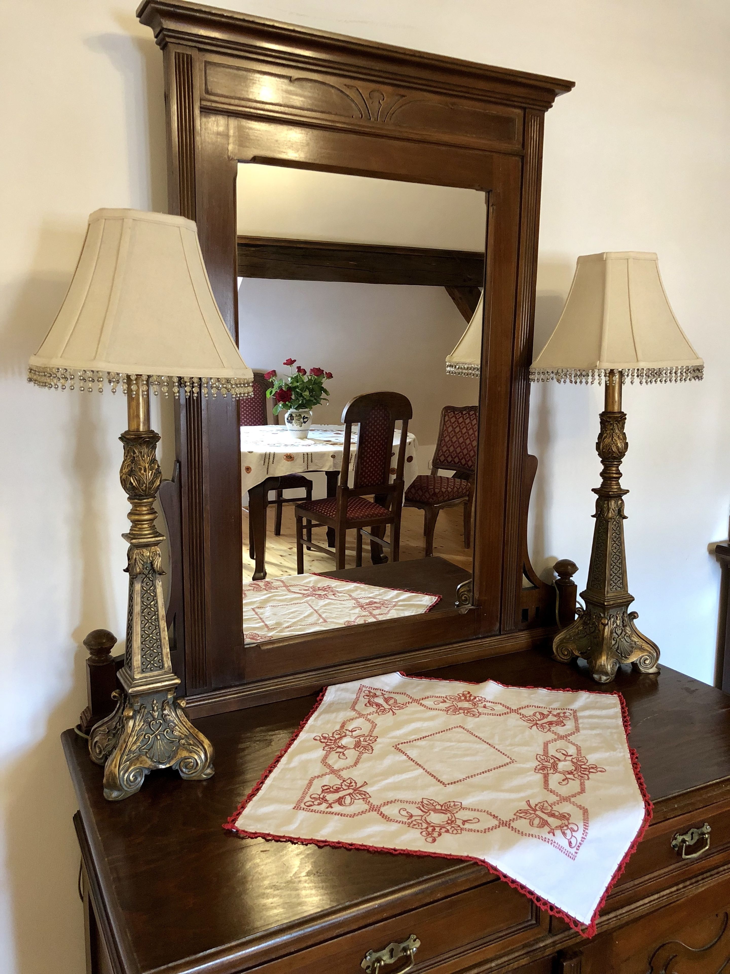 A wooden dresser with two ornate lamps, a mirror reflecting a dining area, and a decorative tablecloth.